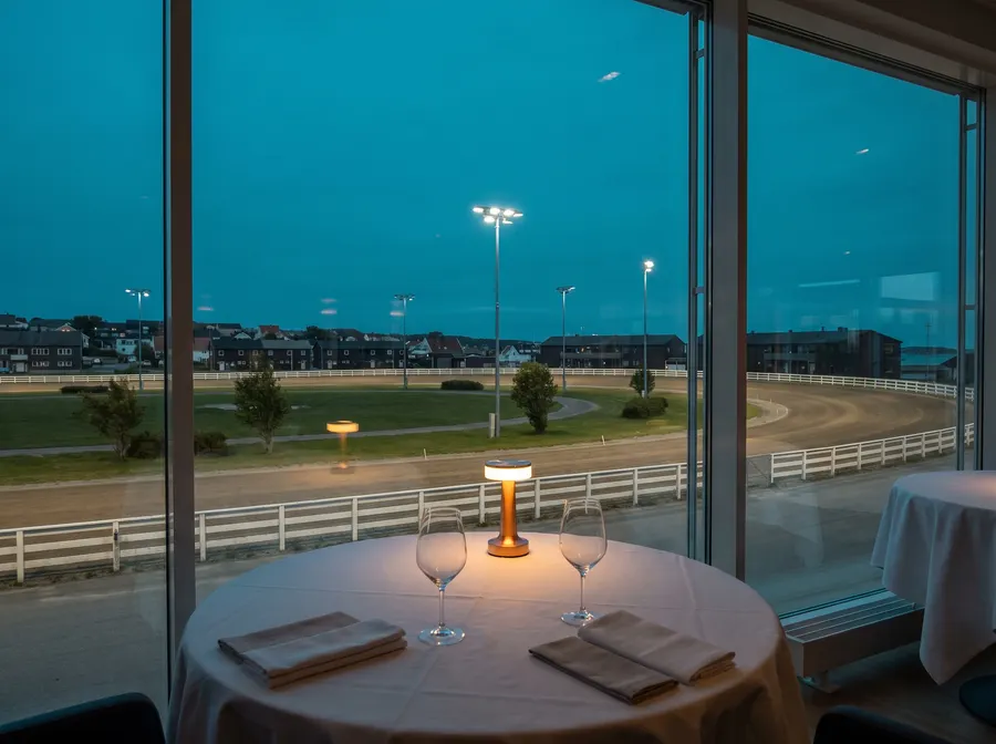 Scène générée : une table pour deux près d'une haute fenêtre à l'heure bleue, avec au loin une piste ovale en terre éclairée par des projecteurs; il ne s'agit pas d'une photo du restaurant.